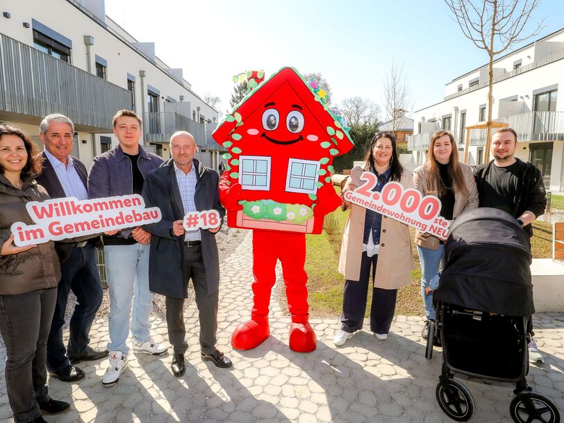 Seven people stand in the courtyard of a residential building. The facility is new and has three floors. In the middle of the group stands the mascot of Viennese housing construction, Wohna Lisa. It is a red friendly looking house with a gable roof on two legs. The people hold the slogans 'Welcome to the municipal building', '#18' and '2,000th municipal apartment NEW' in the hands. A stroller stands in front of two people.