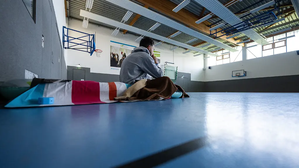 Student sitting in a gymnasium on a sleeping mat representing emergency housing conditions in Münster