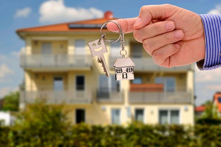 A hand holding a key with a house-shaped keychain against a modern background, symbolizing property ownership