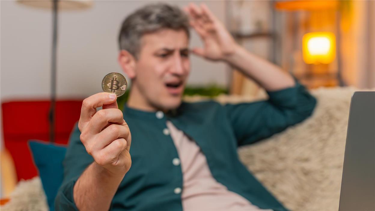 A man holding a Bitcoin coin to the camera, pulling his hair in frustration.