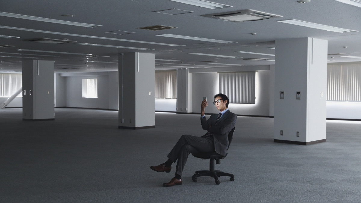 A man in a suit sits alone in an empty office, looking at his smartphone in a moment of financial stress.