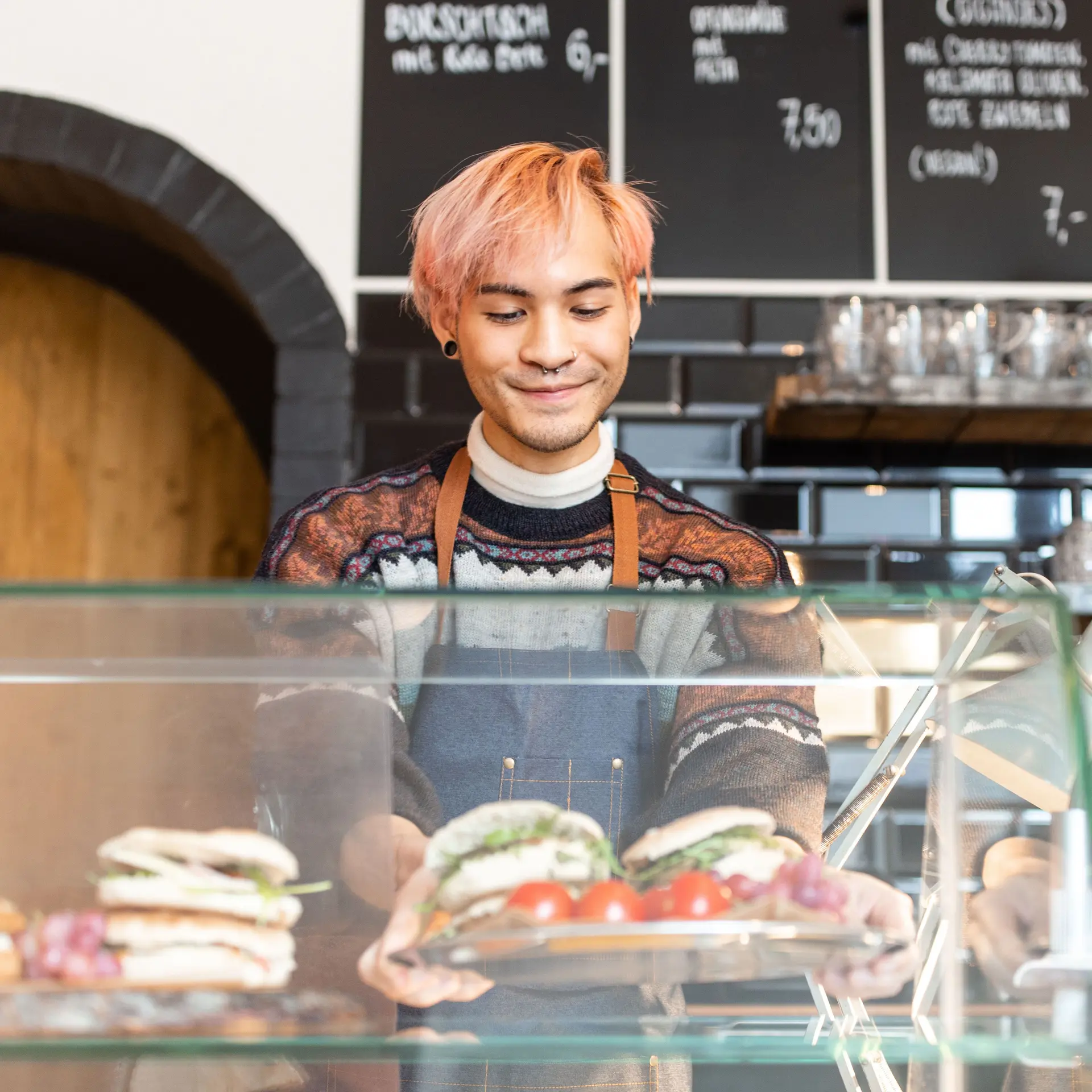 Ein Mann mit rosa gefärbten Haaren arbeitet in einem Cafe und befüllt die Theke mit Paninis.